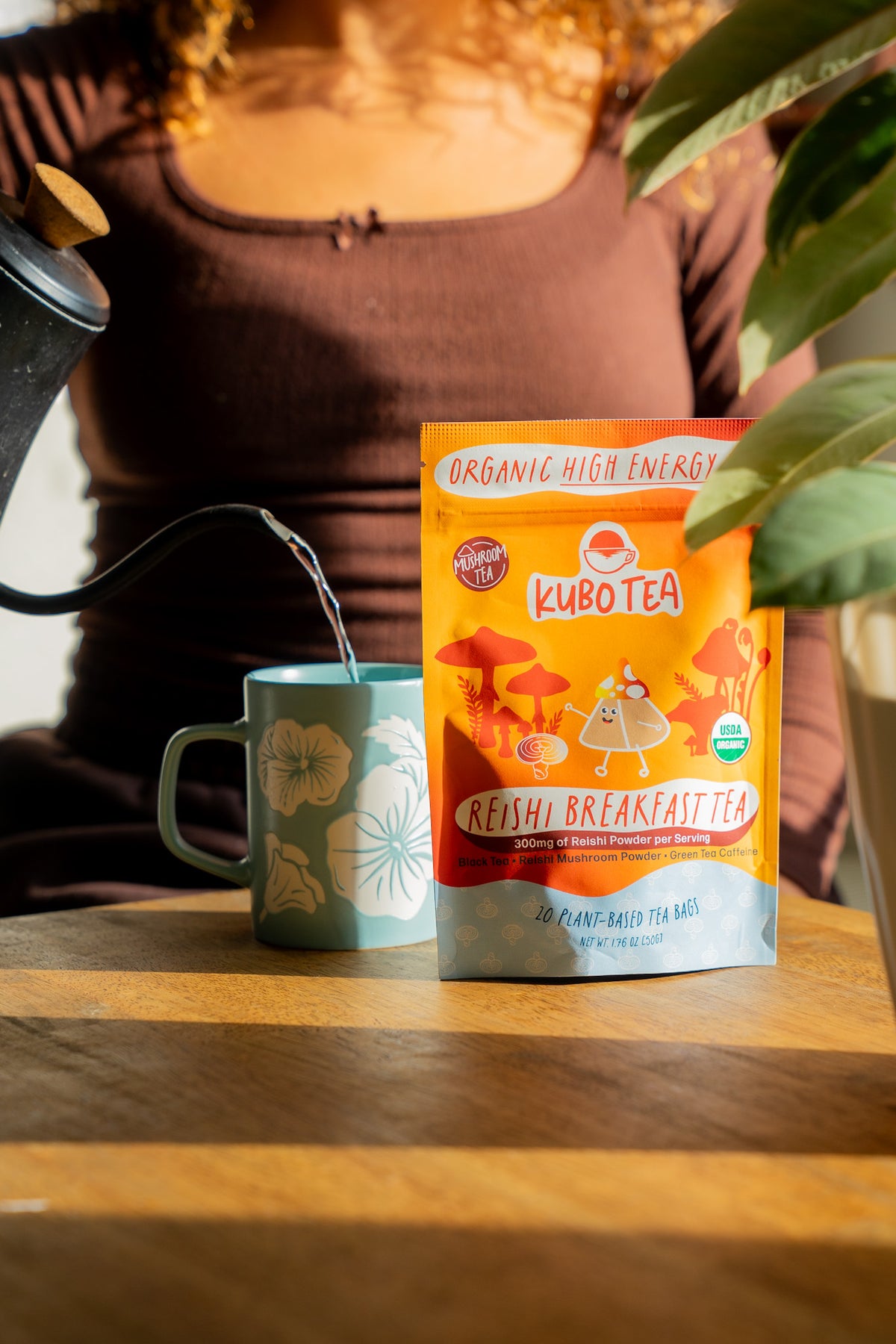 A pouch of Kubotea Reishi Breakfast Tea stands on a wooden table next to a teal mug with a floral pattern, with hot water being poured into the mug from a black kettle. A person wearing a brown top is blurred in the background, and a green plant leaf is visible in the foreground.