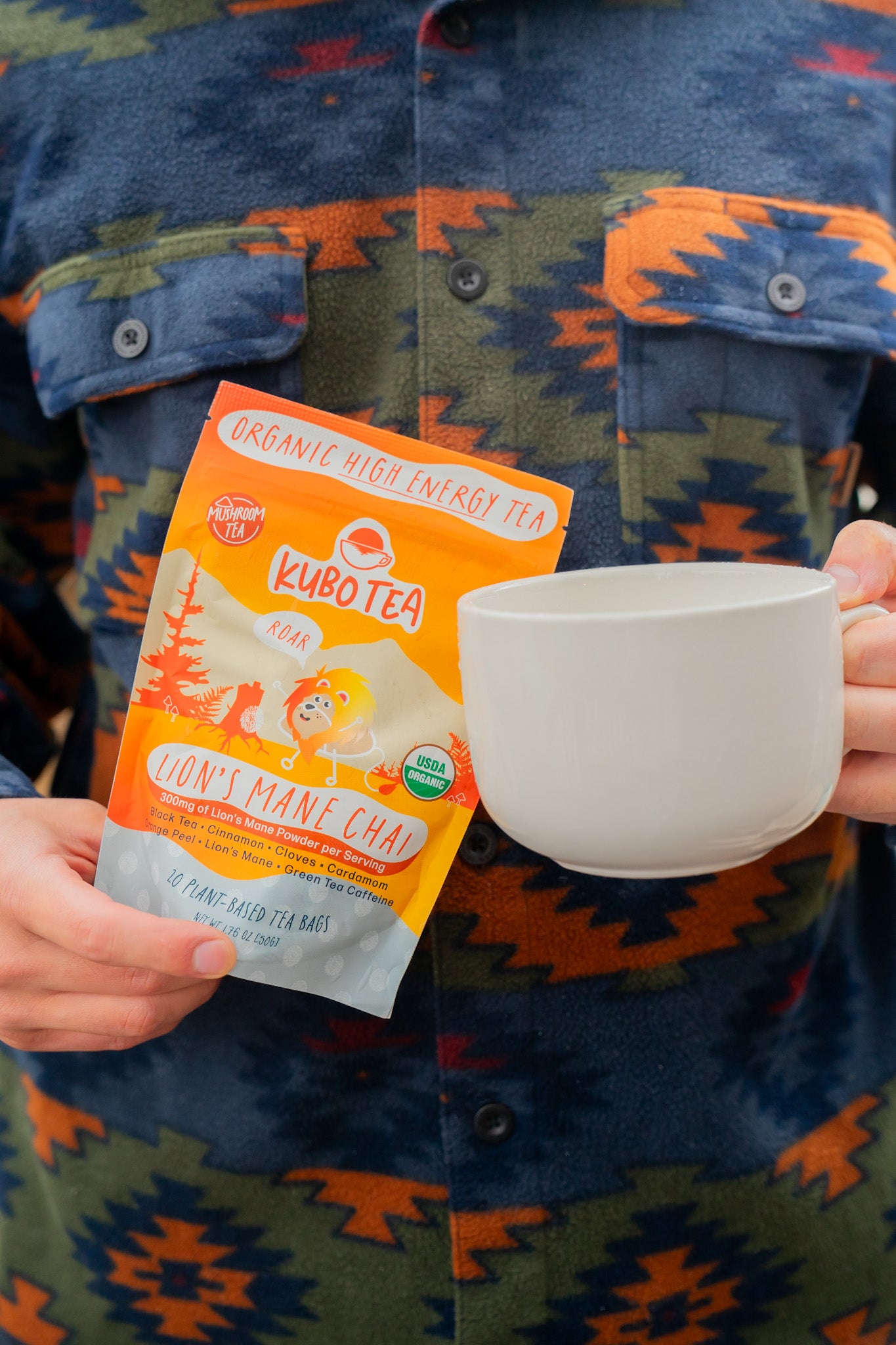 A person wearing a blue, orange, and green patterned shirt holds a packet of Kubotea Don't Mine Chai tea next to a white ceramic mug.