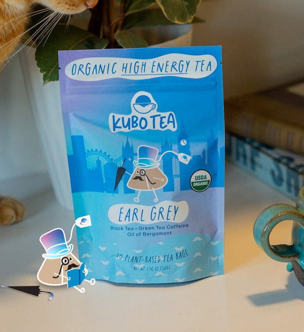 A blue pouch of Kubo Tea Earl Grey organic high energy tea stands on a tabletop in a real indoor setting, with a ceramic mug, books, a plant, and part of a cat visible around it. A small cartoon top-hat character graphic appears near the bottom left.