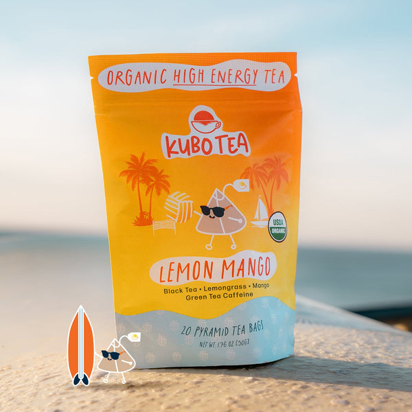 A stand-up pouch of Kubo Tea Lemon Mango organic high energy tea stands on a sandy surface outdoors with a blurred beach or ocean background. The package is bright yellow-orange with a light blue lower section and printed illustrations of palm trees, a drink, and a small cartoon character.