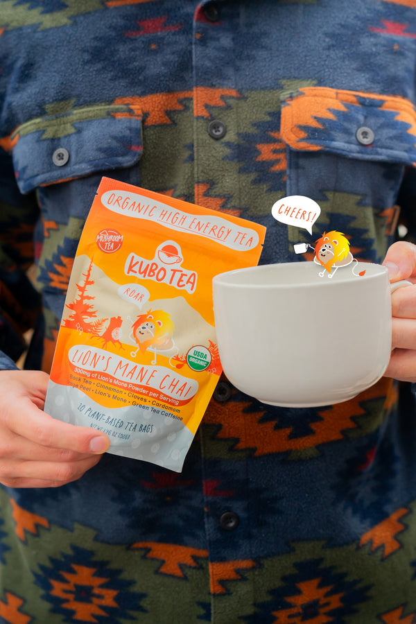 A person wearing a blue and orange patterned button-up shirt holds a bag of Kubotea Organic High Energy Tea and a white ceramic mug. The tea bag packaging is orange and white with cartoon graphics, and a small cartoon character is clipped to the side of the mug.