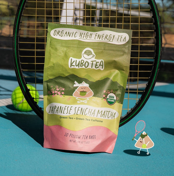 A bag of Kubo Tea Japanese Sencha Matcha tea stands upright on a blue tennis court in front of a tennis net, with a tennis racket behind it and tennis balls around it.
