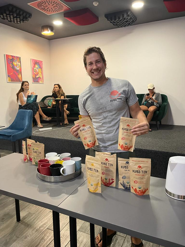 A smiling man in a gray t-shirt stands behind a table holding two bags of Kuebi Tea, with several other tea bags and mugs displayed on the table in a modern indoor lounge setting.
