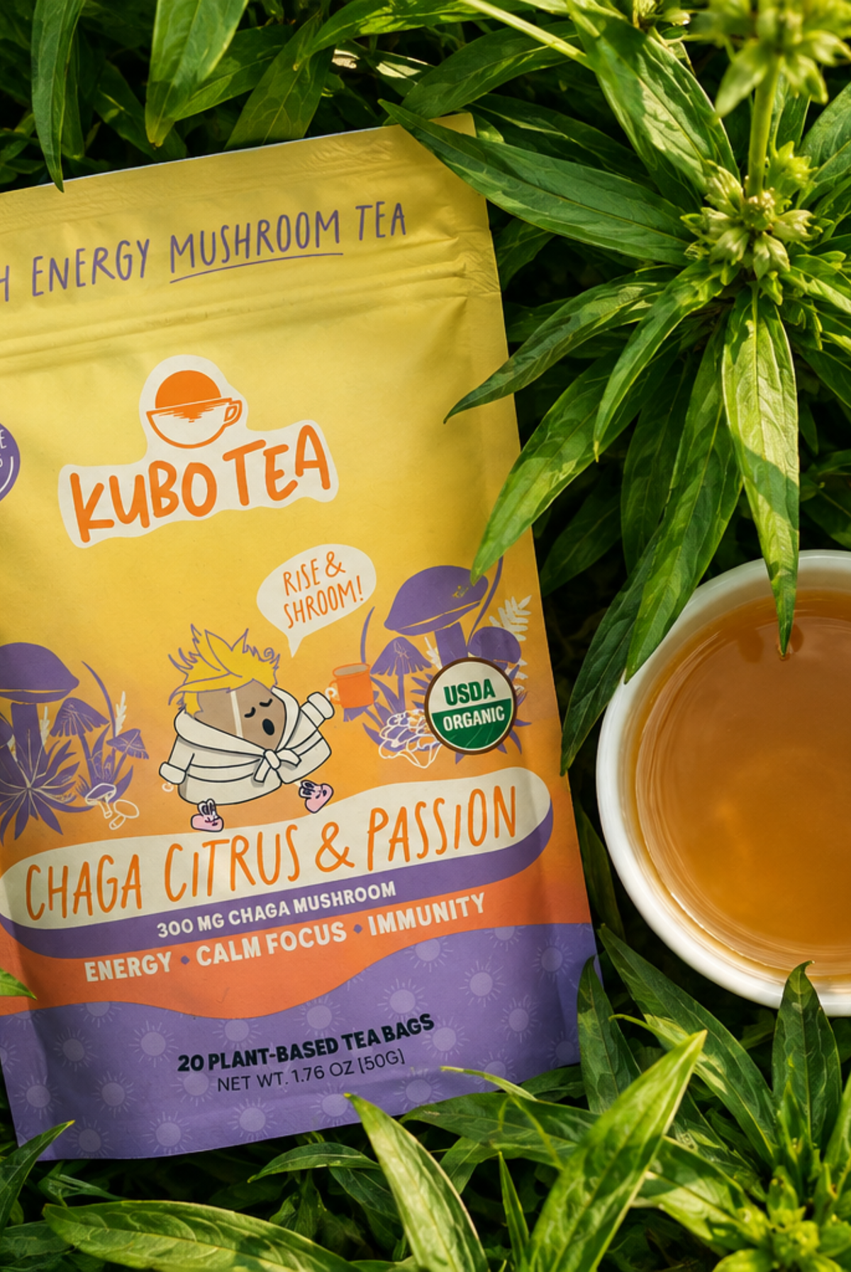 A bag of Kubo Tea Chaga Citrus & Passion Energy Mushroom Tea is displayed among green leaves next to a white cup of brewed tea.