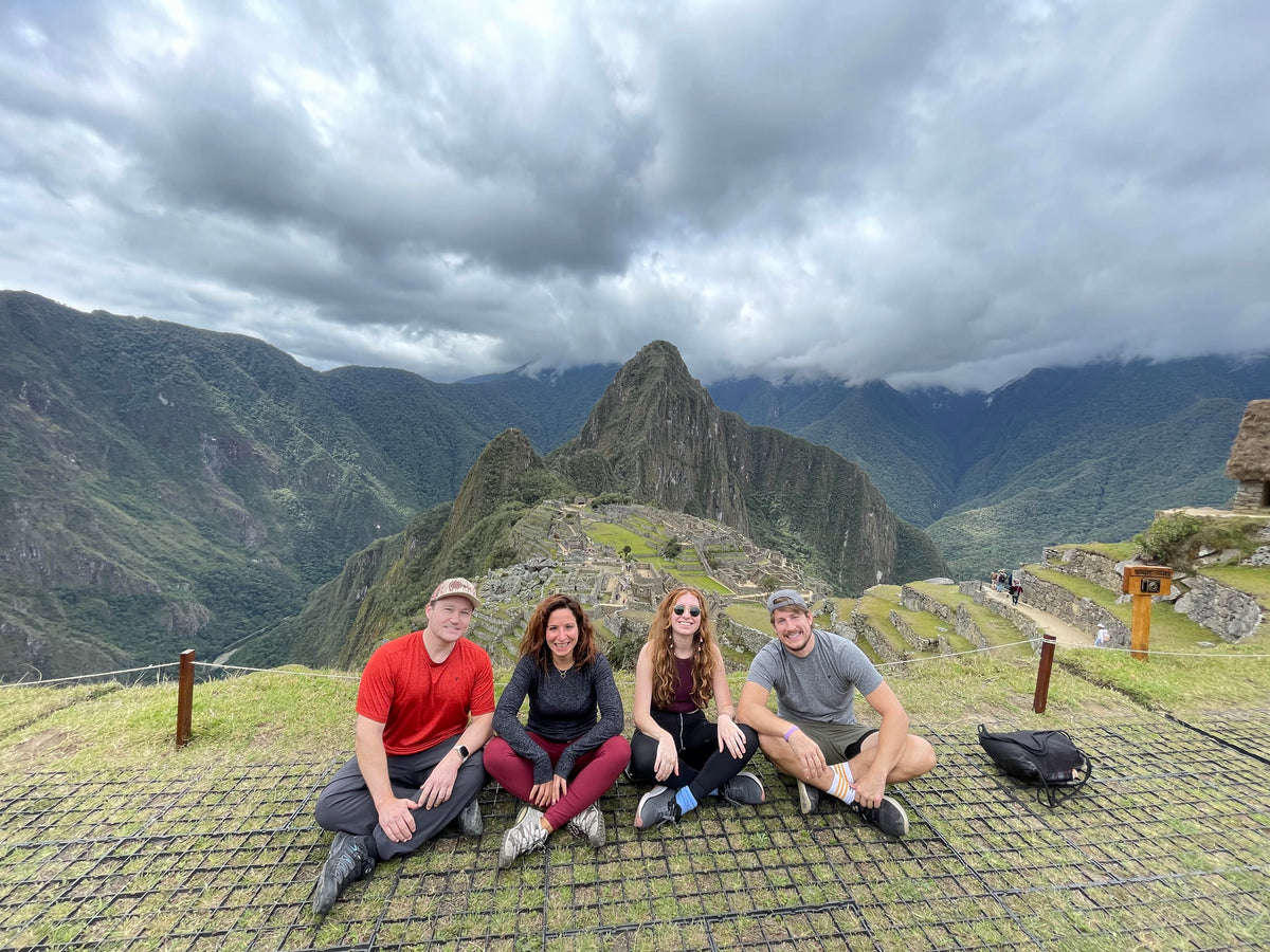 Four friends sitting together on a stone terrace at Machu Picchu, with the ancient ruins and green mountains in the background under a cloudy sky.